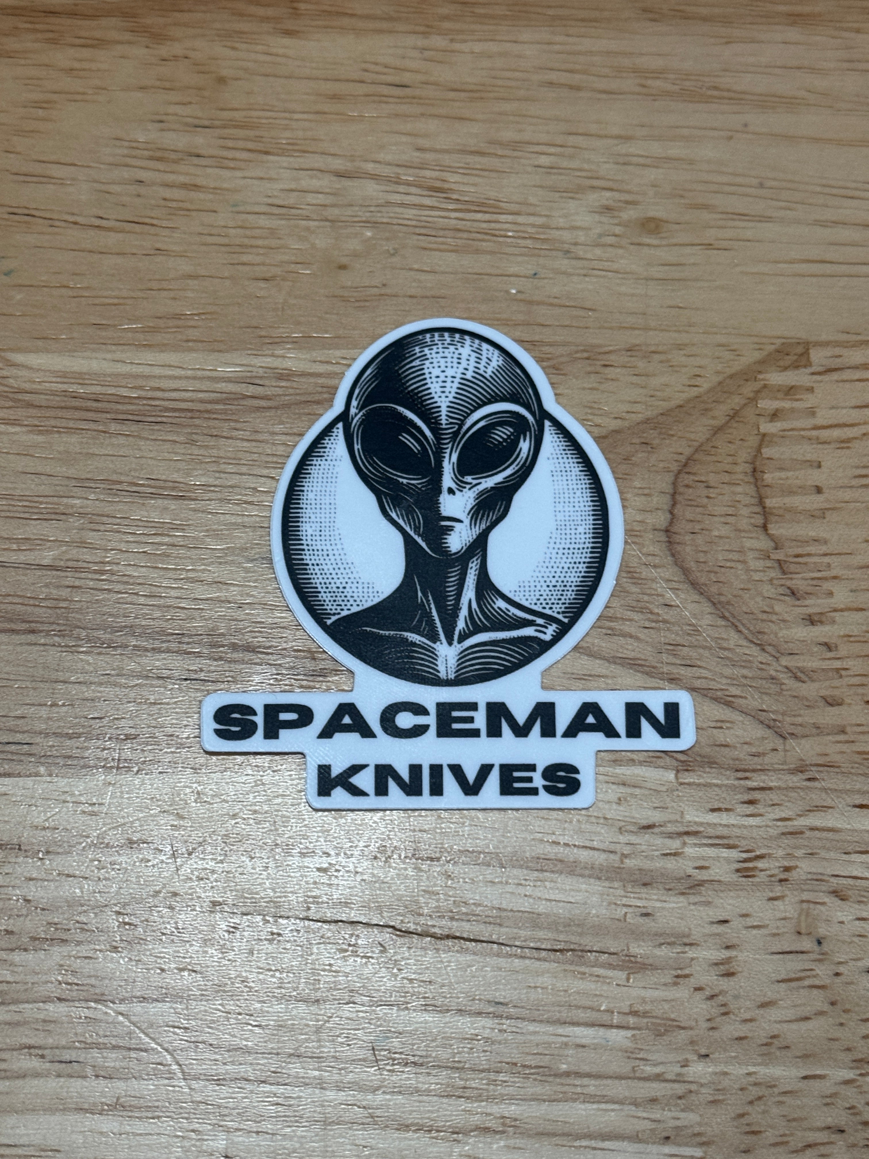 Sticker with alien design and 'Spaceman Knives' text on a wooden surface